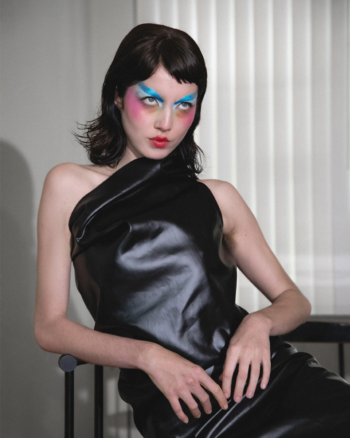 Autry seated in a black leather dress with colorful makeup looking intently past the camera